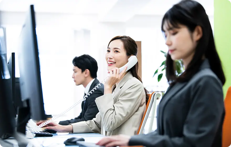 オフィスで3人のビジネスパーソンがパソコン作業をしており、中央の女性が電話対応中